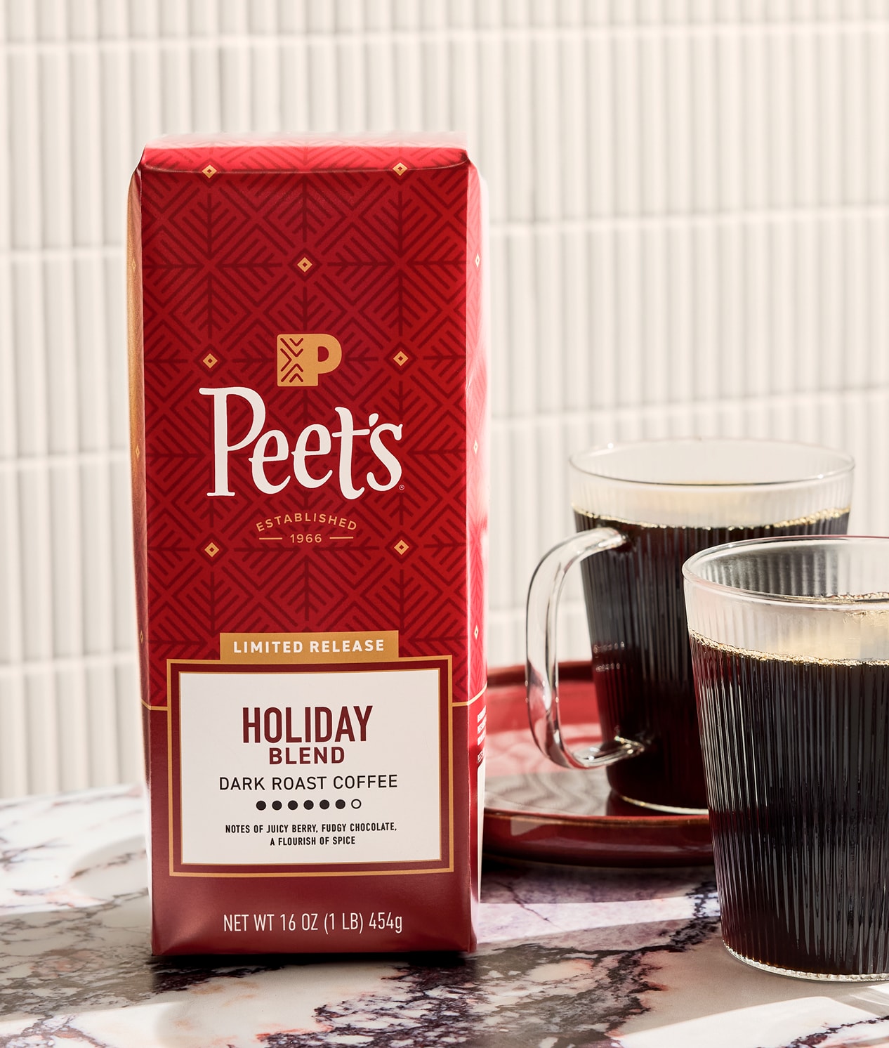A red bag of Peet's Holiday Blend Dark Roast Coffee stands on a marble surface next to two clear glasses of black coffee, with a white tiled wall in the background.
